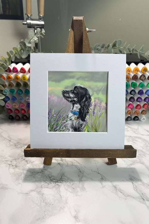 A small framed artwork on a wooden easel, showing a painted black-and-white dog in a grassy field. The frame is white and displayed against a marble-patterned background with colored markers visible behind it.