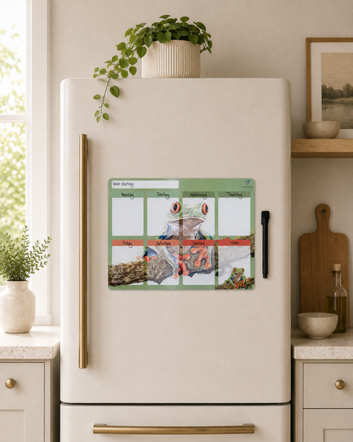 A weekly magnetic calendar planner with a colorful frog illustration is shown on the front of a cream-colored refrigerator in a modern kitchen setting.